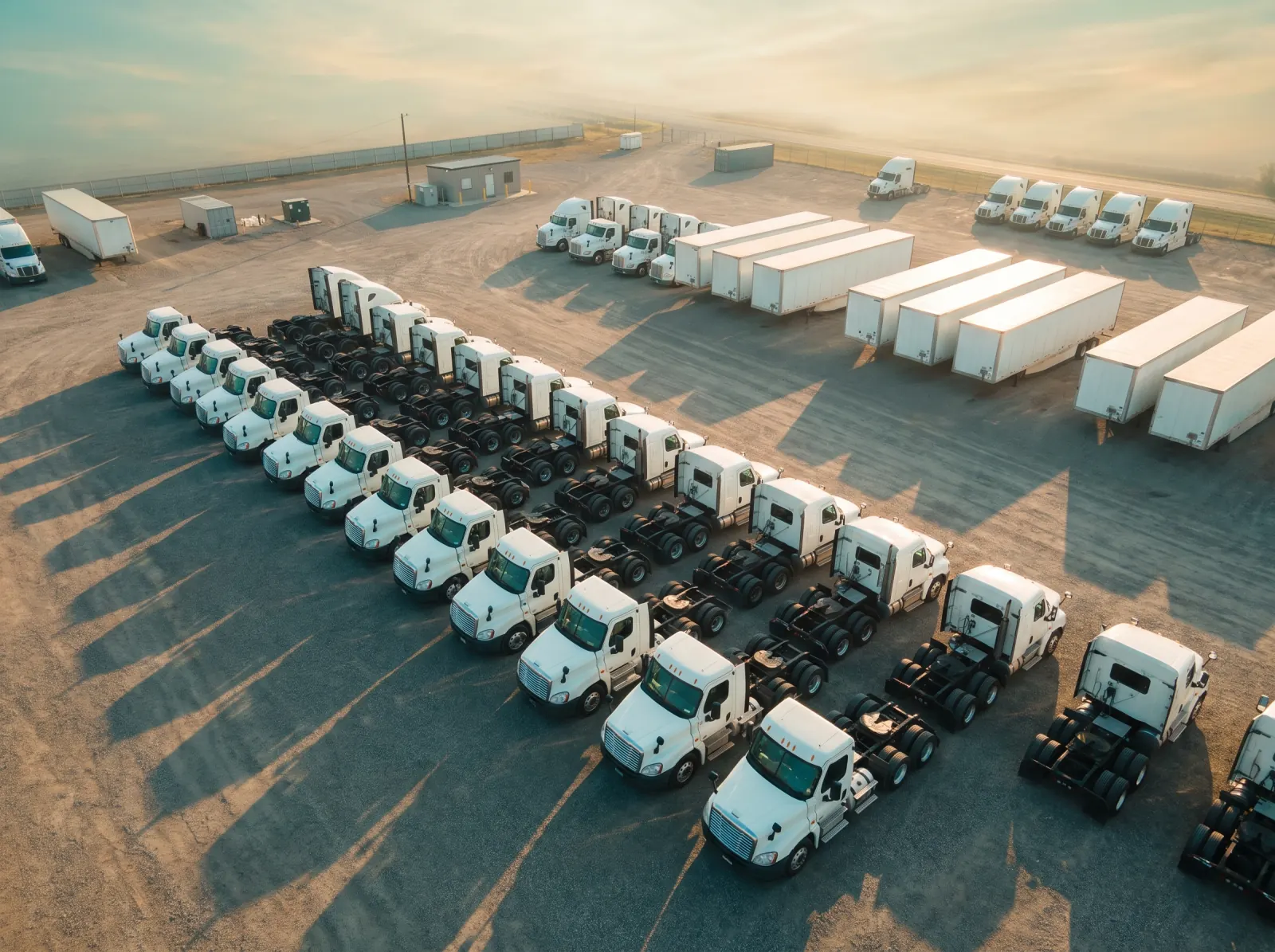 Rows of white semi tractors and trailers parked in a fleet yard, seen from above in low sun.